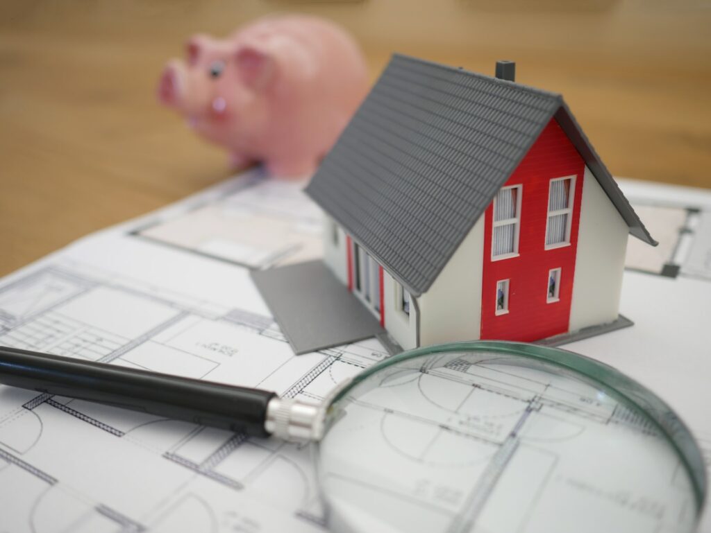 an image of a house blueprint with a piggy bank, magnifying glass, and model house on top of the blueprint
